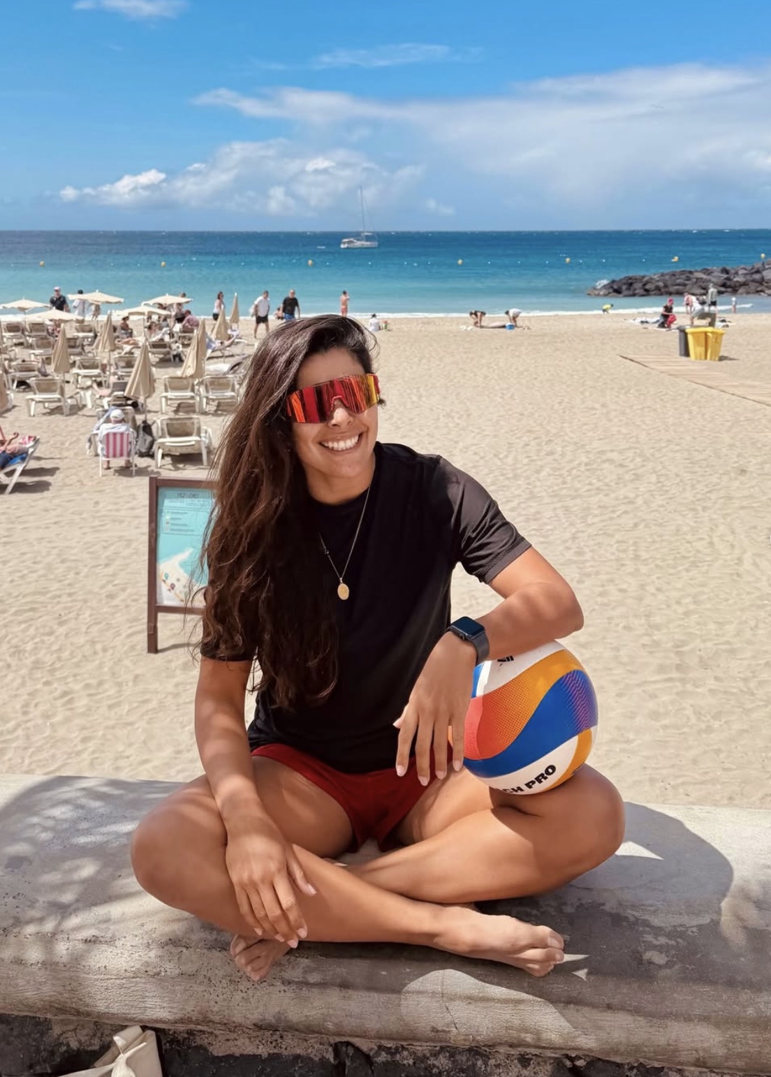 Gaby, founder of Beach Volley Mancora, sitting on a wall with a volleyball, beach behind her
