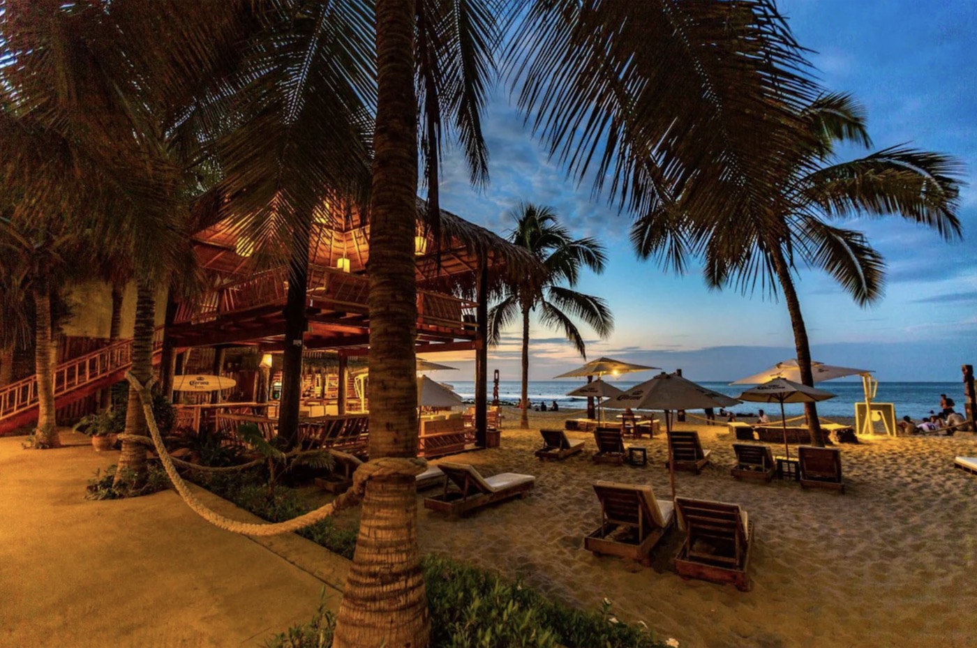 Del Wawa lit up at dusk, with loungers on the sand and the sea beyond